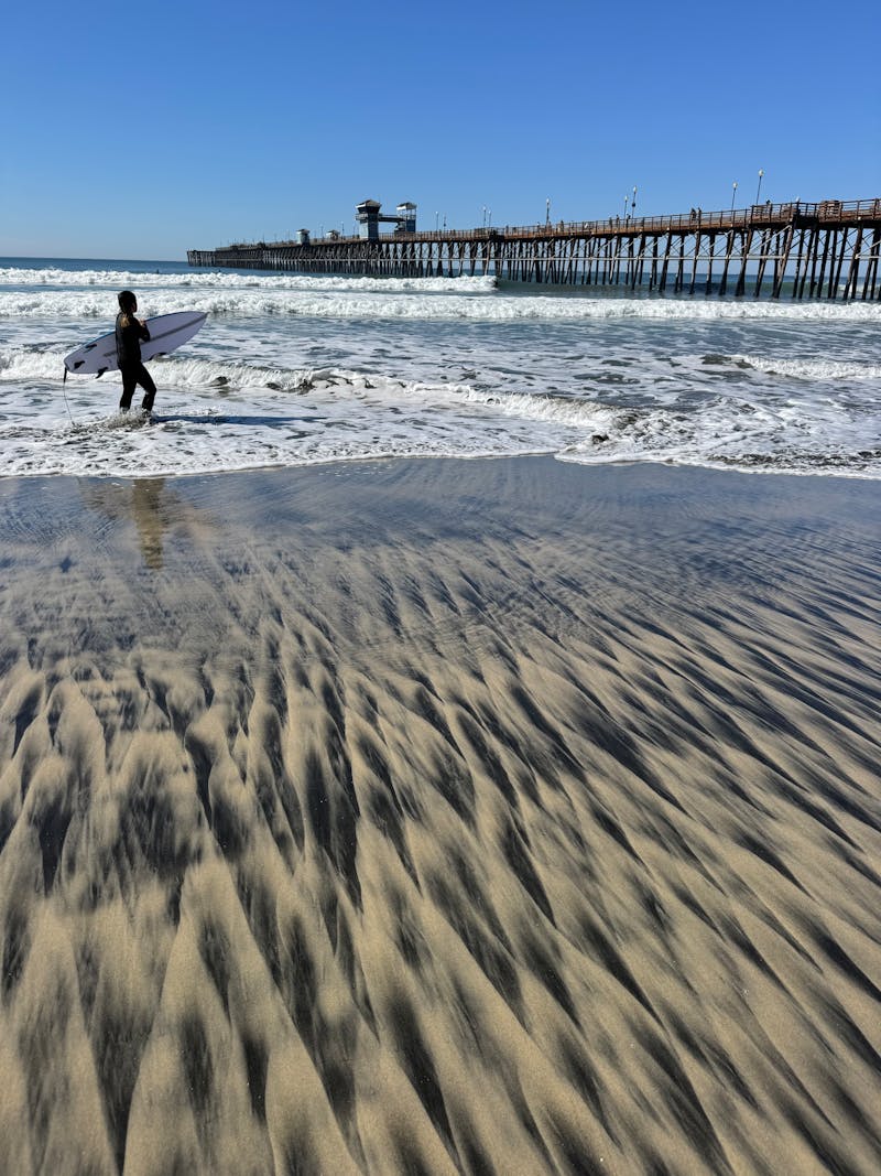 A surfer walks along the shore near Oceanside Pier